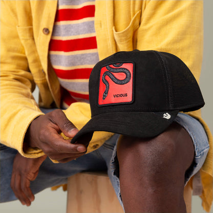 Wearing a yellow jacket and striped shirt, a person holds a Goorin Bros. Serpente Rosso black microsuede cap with a red snake patch, resting it on their knee.
