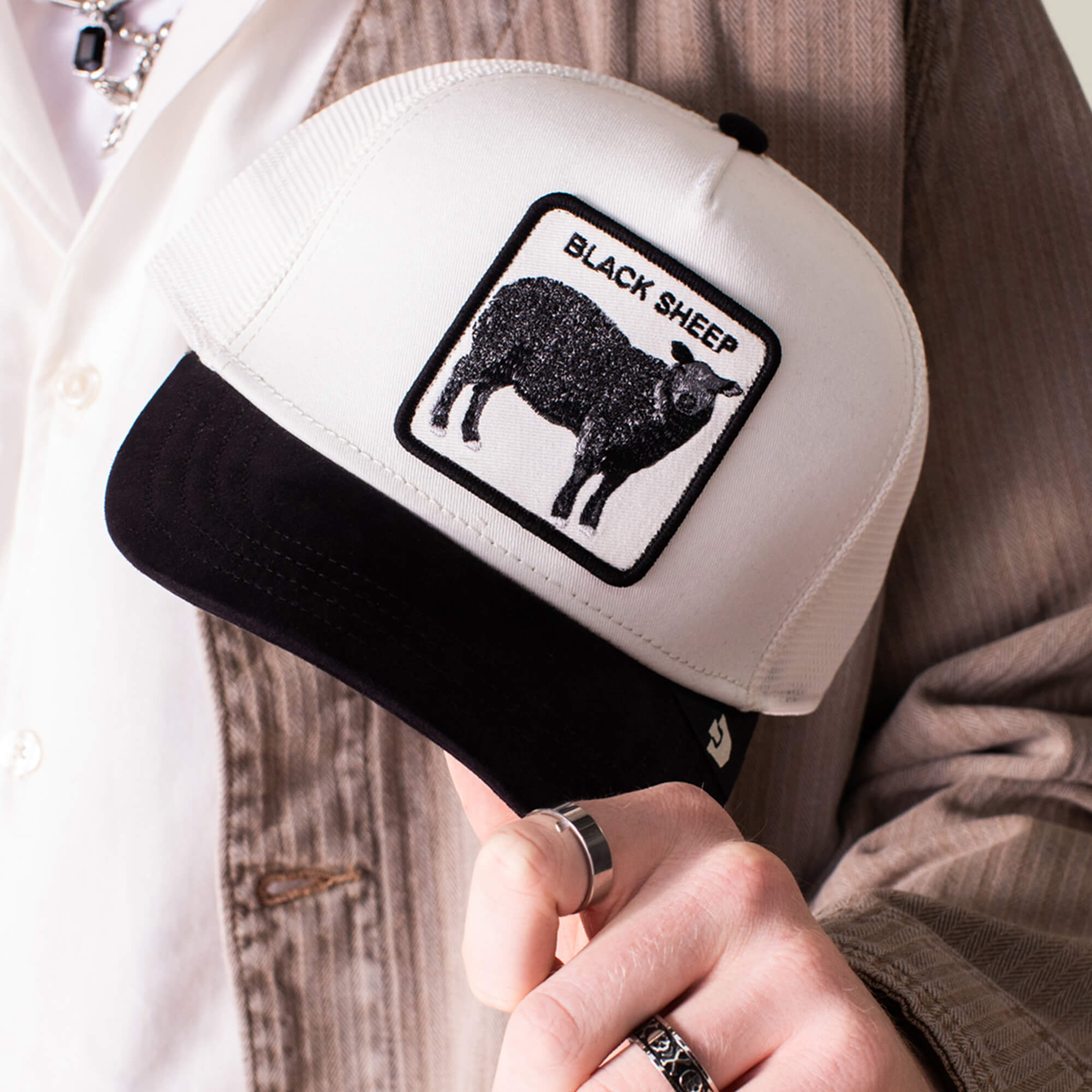 A person holds the Suede Sheep cap by Goorin Bros.—a black and white hat with a suede brim, featuring a black sheep patch and the text BLACK SHEEP.