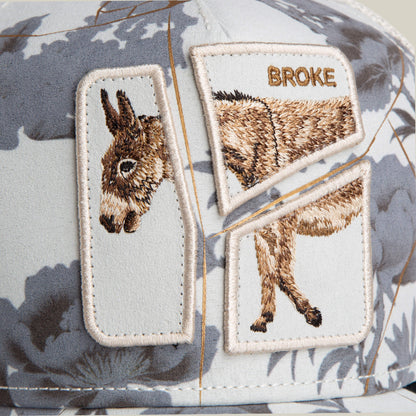 The Goorin Bros. "Break The Bank" cap features embroidered patches with a broken donkey image and "BROKE" above it, set against a gray and white abstract background—an ideal novelty gift for anyone short on cash.
