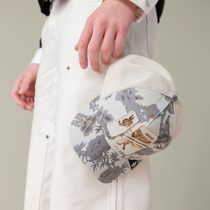 A person in white pants holds the Goorin Bros. "Break The Bank" baseball cap, a light floral-patterned hat with an embroidered cat design and rectangular tag—a standout novelty gift.
