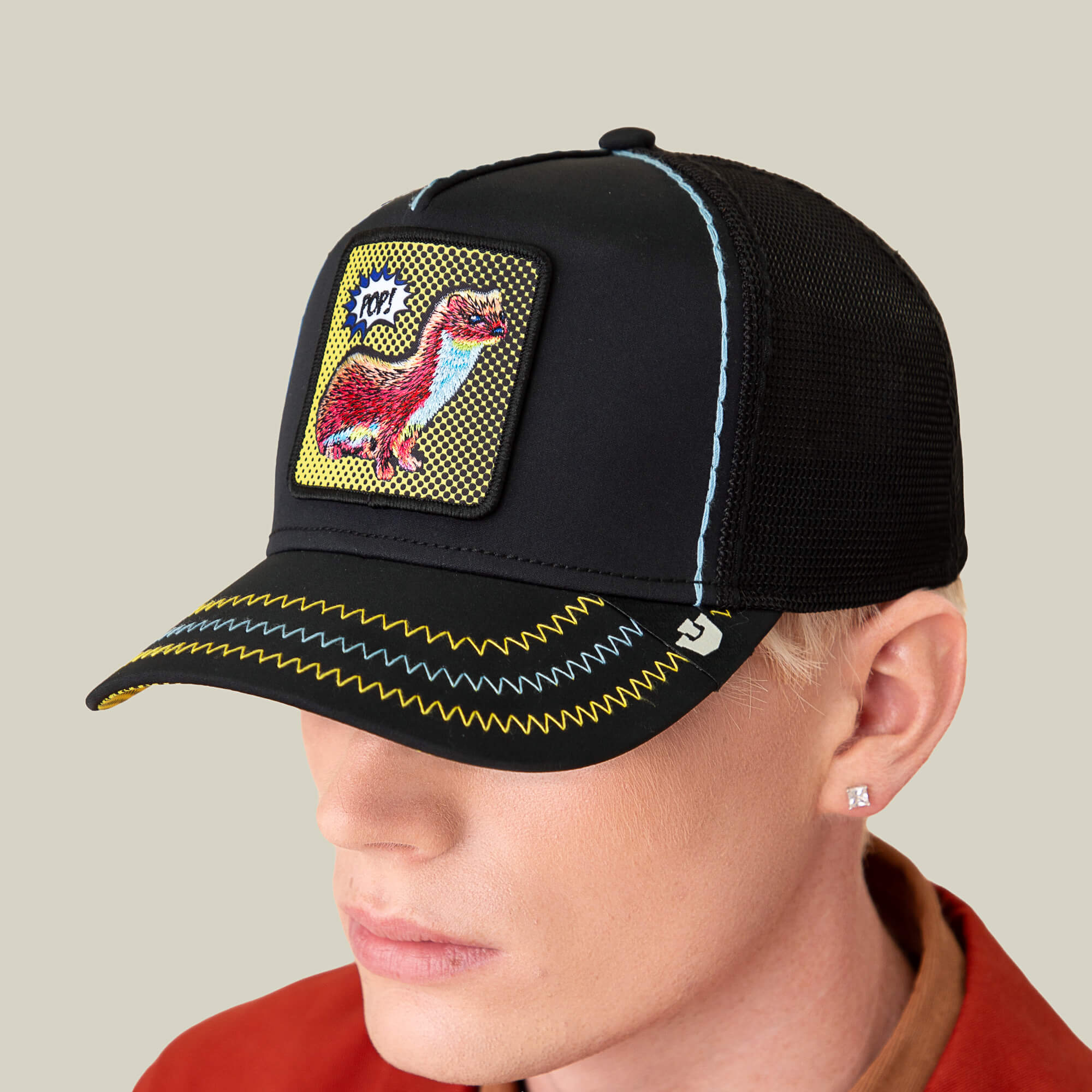 A person sports Goorin Bros.' All Around the Mulberry Bush hat with a dinosaur patch and bold yellow stitching, paired with a red-collared shirt, giving off playful vibes against a neutral background.