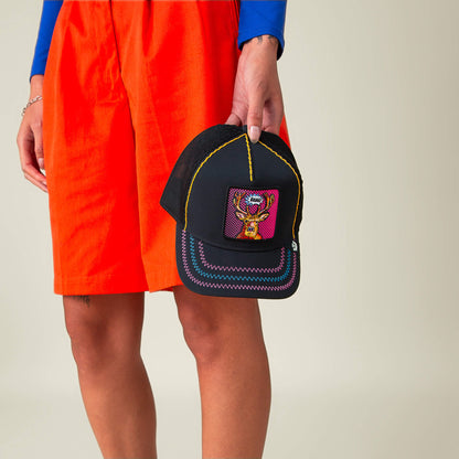 Someone in orange shorts holds the "Earned Media" black cap by Goorin Bros., featuring neon stitching and a colorful Pop Art-inspired embroidered design, set against a plain background.