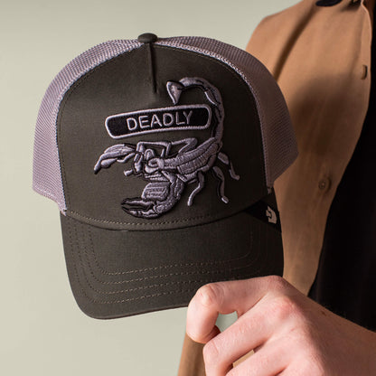 Someone holds the Goorin Bros. Classic Rocker - Deadly trucker hat, which has a military green front with an embroidered scorpion patch and “DEADLY,” plus a grey mesh back.