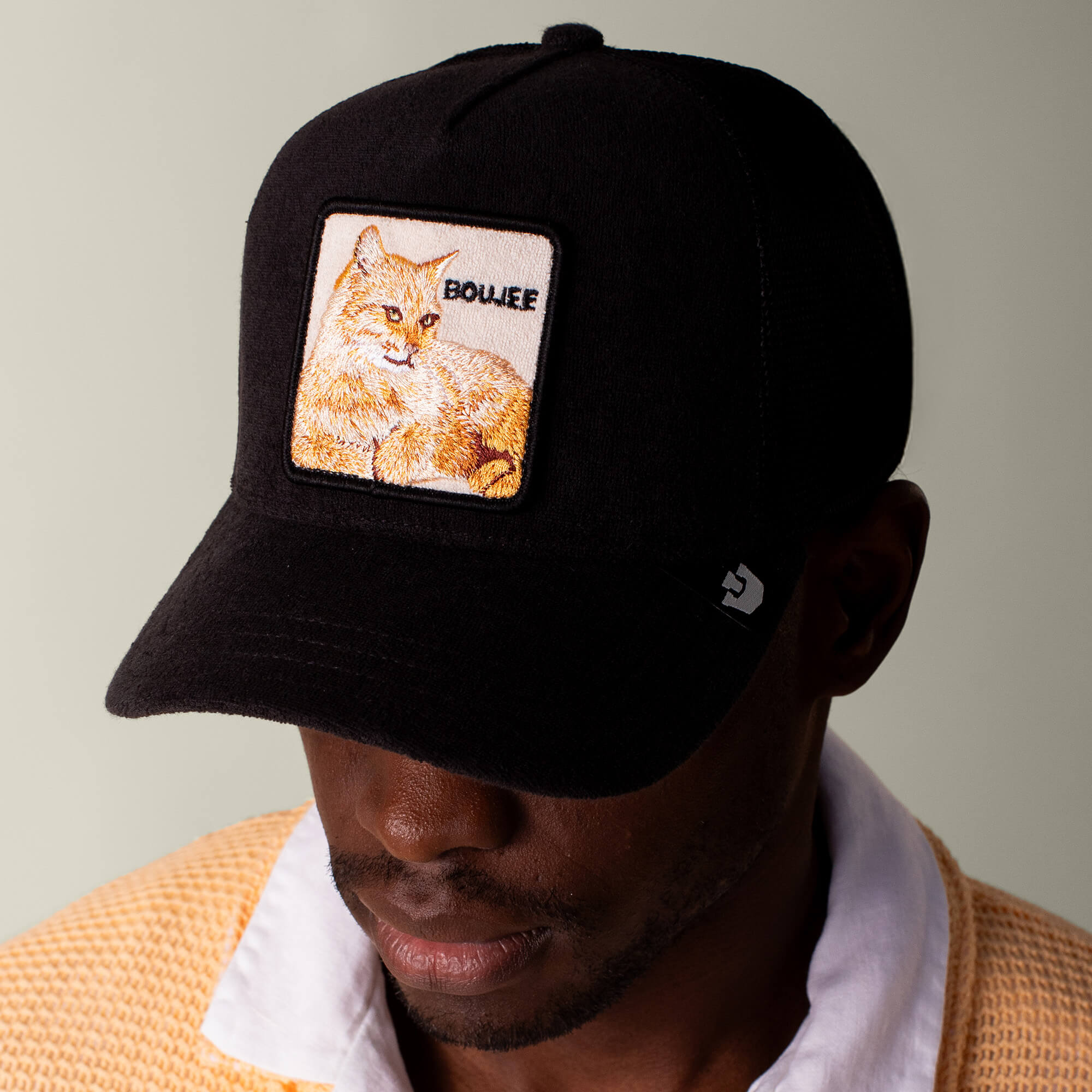 A person sports the Goorin Bros. Terry Cloth Cat cap, a black terry cloth hat with an embroidered cat patch and "DOUZE," paired with a light sweater and white collared shirt for plush lounge-level softness.