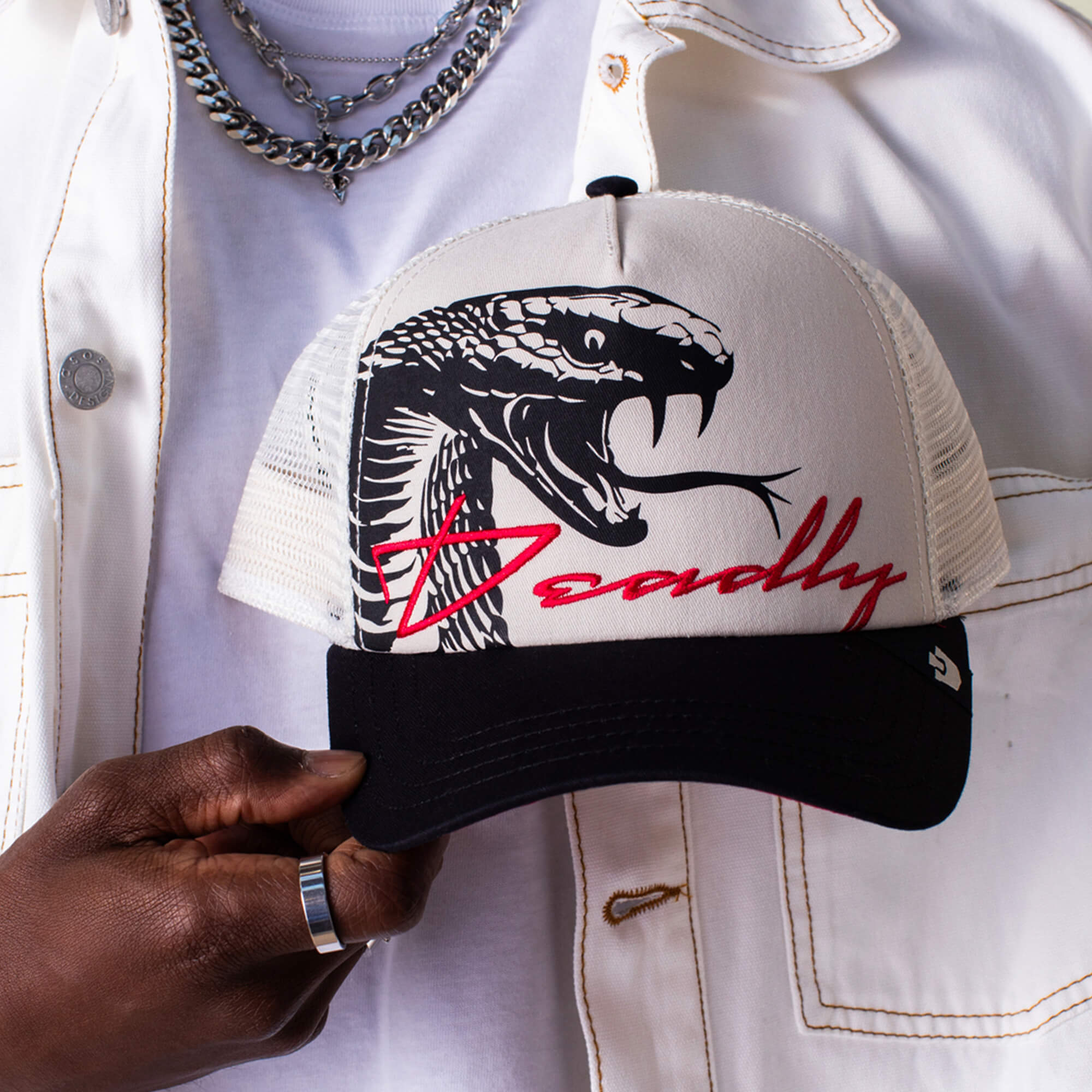 A person in a white jacket holds the Goorin Bros. "Outside the Lines Snake" cream and black trucker hat featuring a snake illustration and "Deadly" in red script.