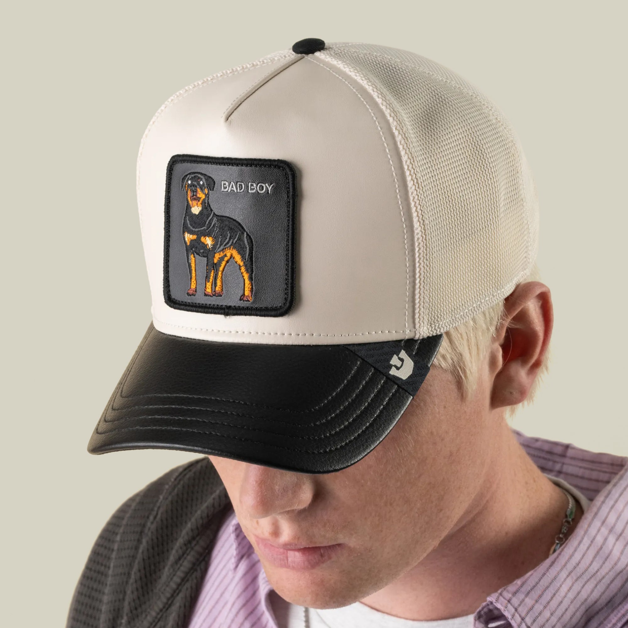 A person wearing the Goorin Bros. Faux Leather Rottweiler trucker cap—black and white faux leather with an embroidered patch featuring a dog and the words “BAD BOY”—is looking down.