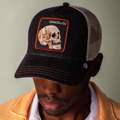 A man looks downward wearing the Goorin Bros. Denim Skull black and white mesh trucker cap, featuring a skull patch with “CANCELLED” embroidered above. He pairs it with a yellow textured shirt and dark wash black denim.