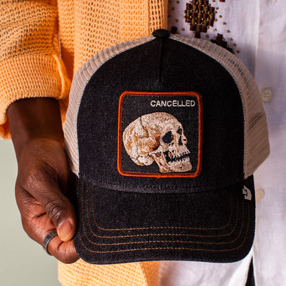A person holds the Goorin Bros. Denim Skull black and white mesh baseball cap, featuring a skull patch labeled "CANCELLED," perfectly styled with dark wash black denim for an edgy look.
