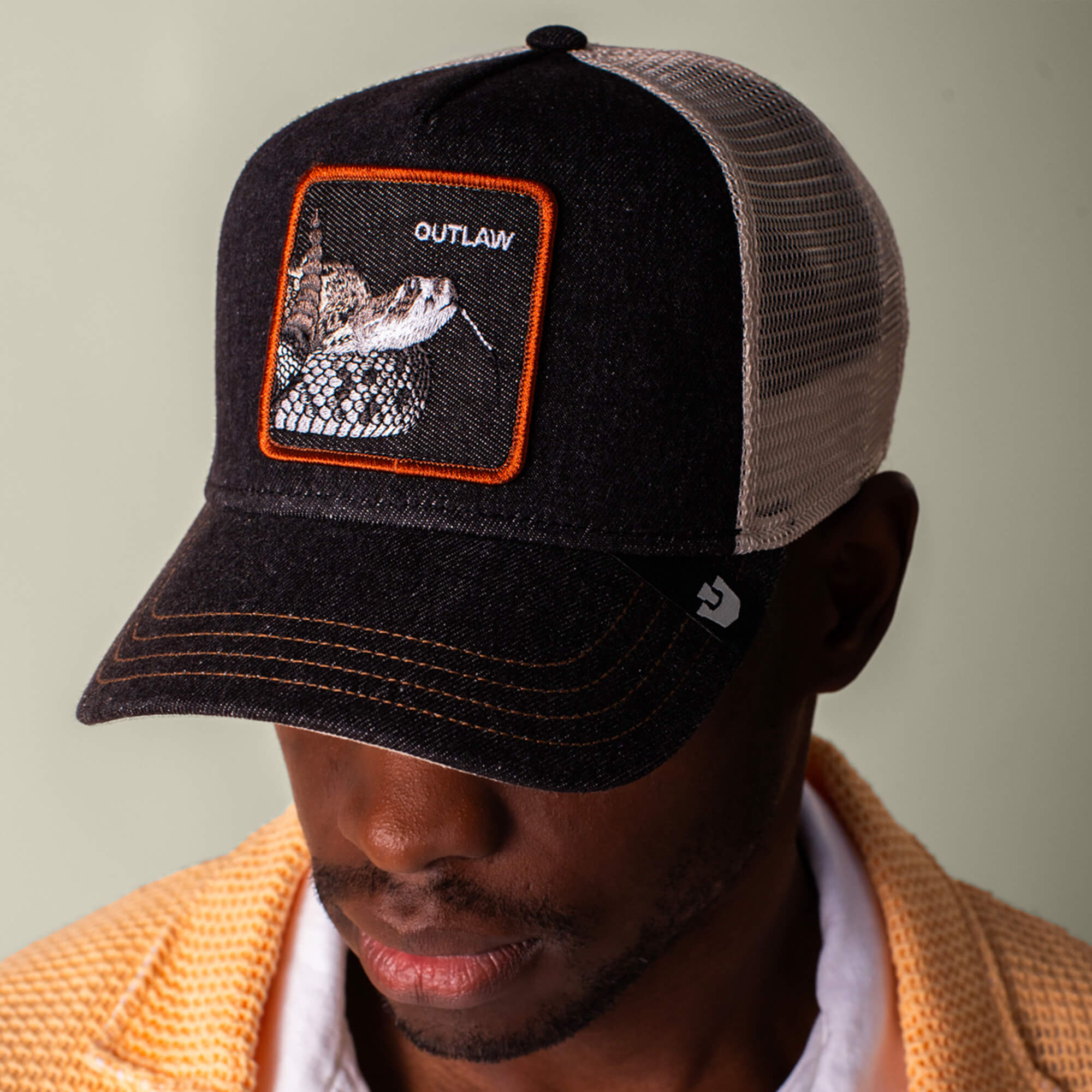 A man sports the Goorin Bros. Denim Snake black and white mesh trucker hat, featuring a bold rattlesnake patch and OUTLAW text, paired with rugged black denim for a rebellious look.