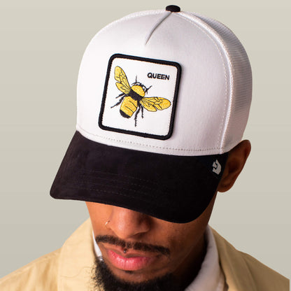 A person wearing the Suede Bee cap by Goorin Bros., featuring a white and black design, a Queen patch with a yellow bee, and a sleek black microsuede brim.