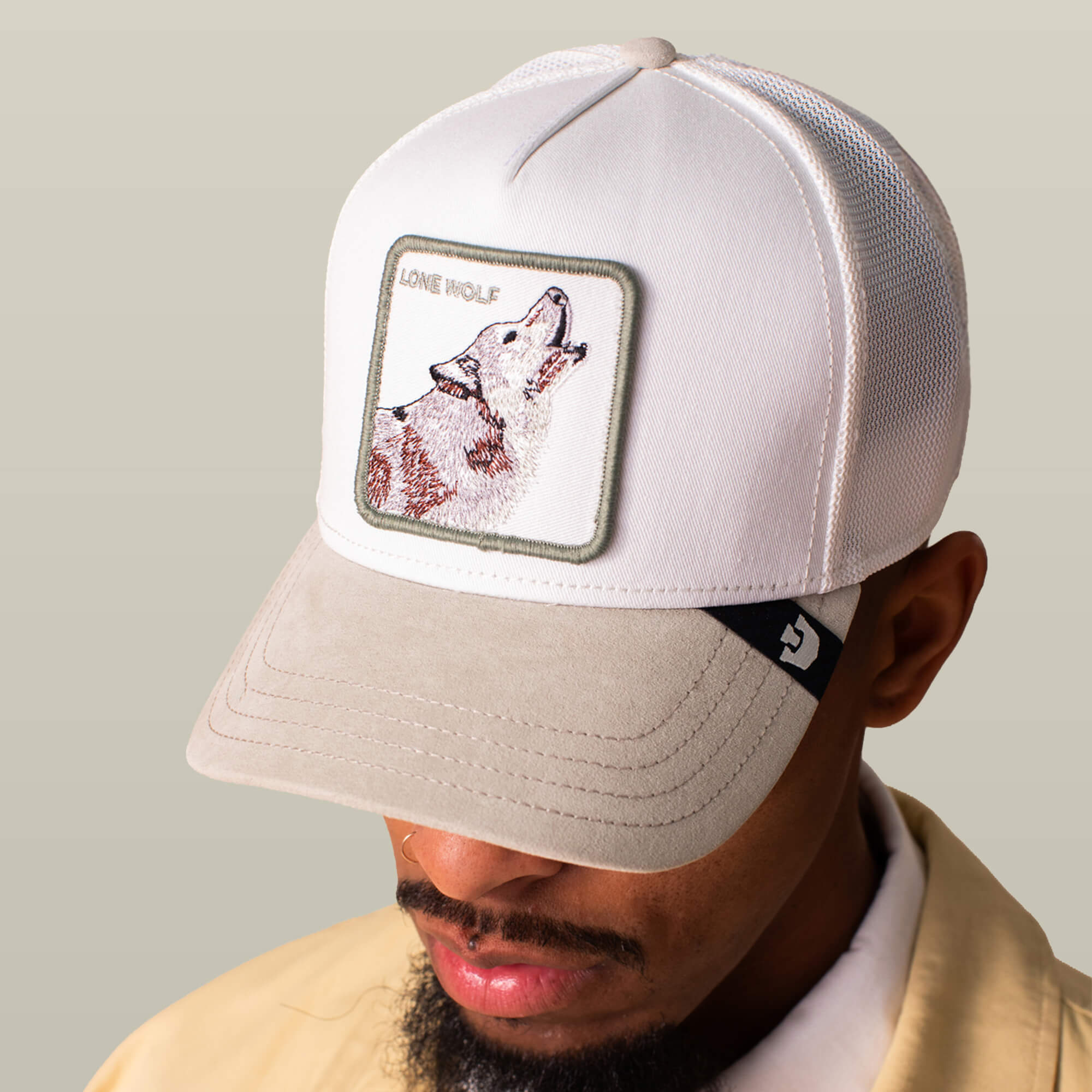 A person wears the Goorin Bros. Suede Wolf trucker hat—white and beige with a suede brim and LONE WOLF wolf patch—while looking down, their face partly hidden.