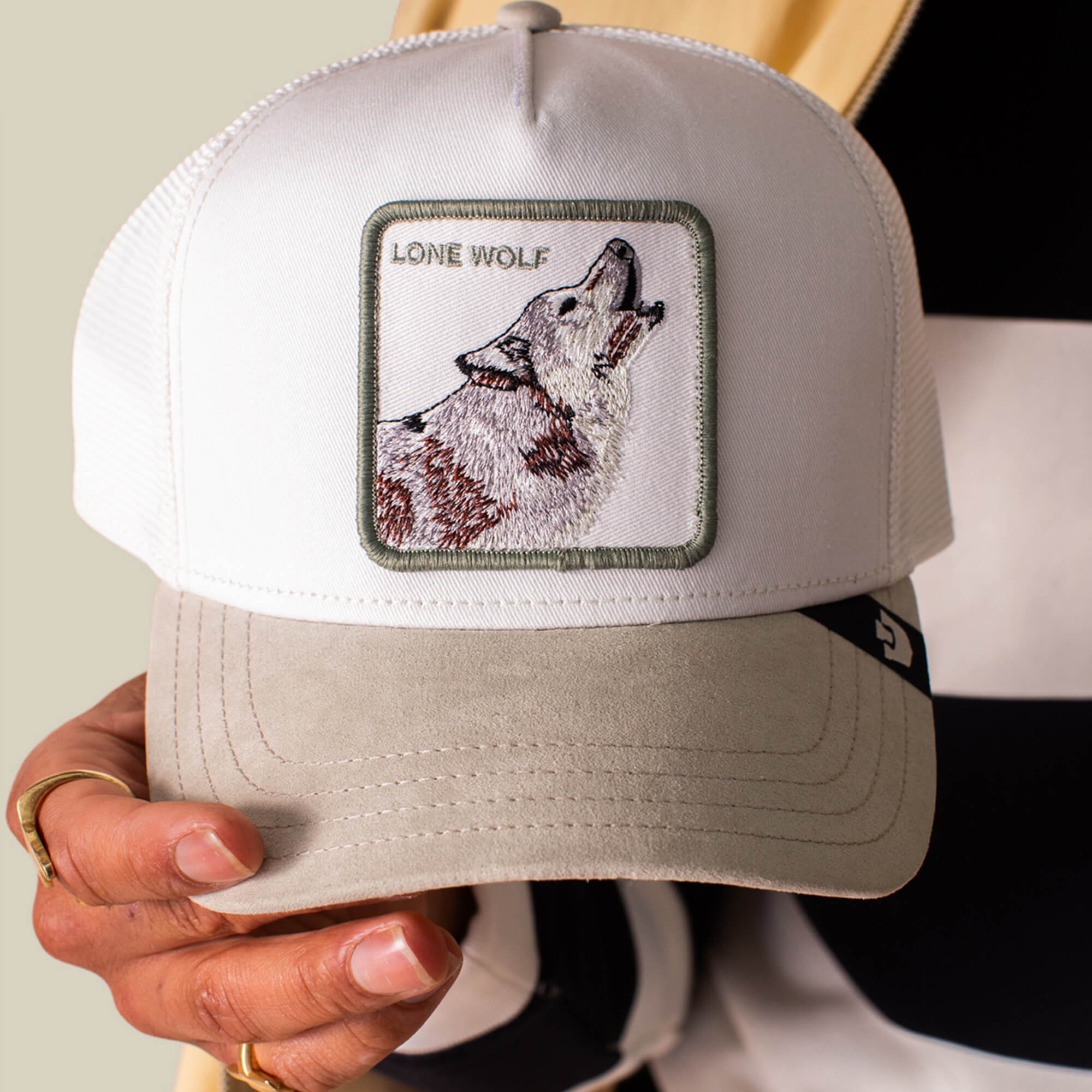 A person holds the Goorin Bros. Suede Wolf trucker hat, white and beige with a suede brim, featuring a wolf patch and "LONE WOLF" embroidered on the front.