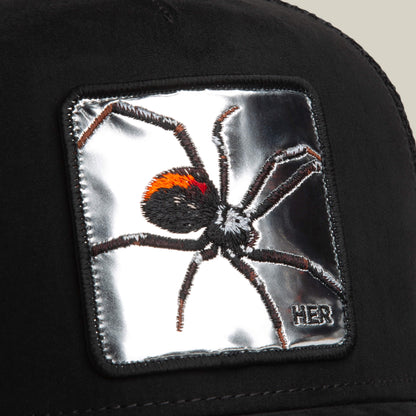 A close-up of the Goorin Bros. Silver Spider trucker hat, showcasing a silver patch with an embroidered black and red spider, plus "HER" stitched in the corner.