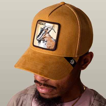 A man wears the Goorin Bros. Golden Goat tan gold suede trucker hat featuring a goat patch and "GOAT" text on the front, set against a plain neutral background that highlights his confident style.