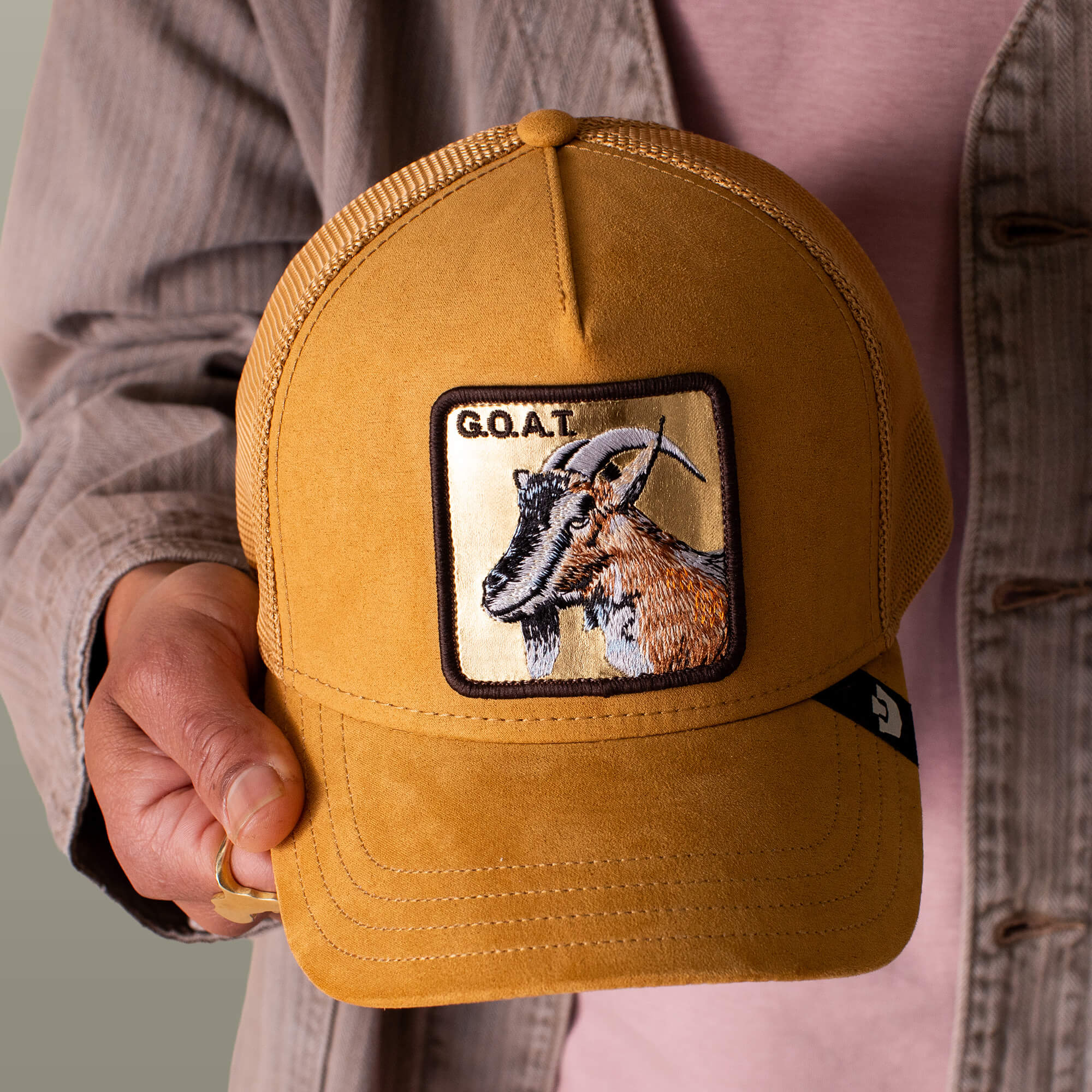 A person holds the Goorin Bros. Golden Goat trucker cap, featuring gold suede and a goat patch embroidered with "G.O.A.T.