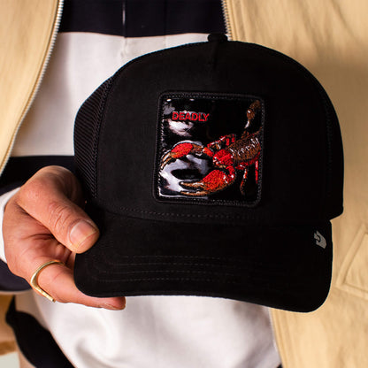 A person holds the Goorin Bros. Black Out Scorpion cap, a sleek black suede baseball hat featuring a scorpion patch with "DEADLY" embroidered above.