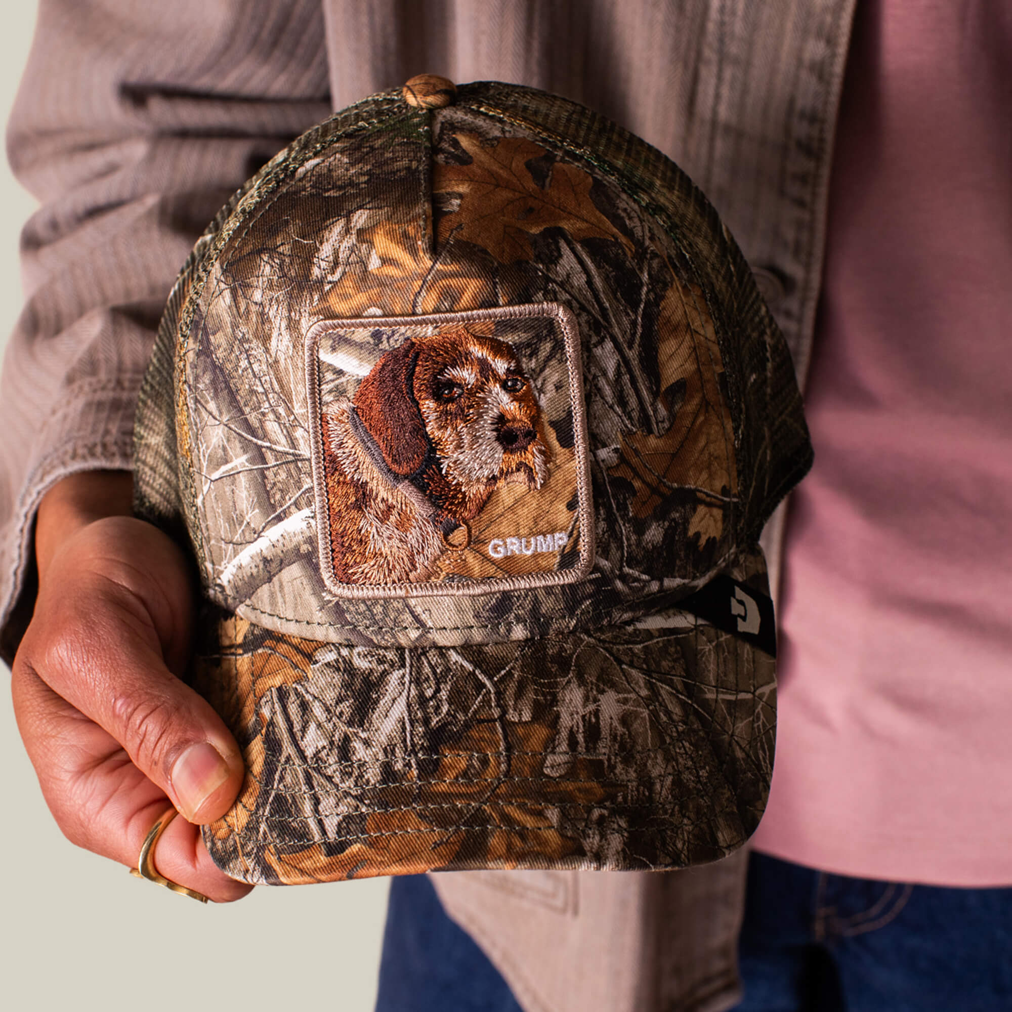 A person holds the Goorin Bros. Realtree Edge Grump Dog cap, showcasing a camo pattern, dog patch, and GRUMP embroidery on the front—perfect for channeling true grump energy.