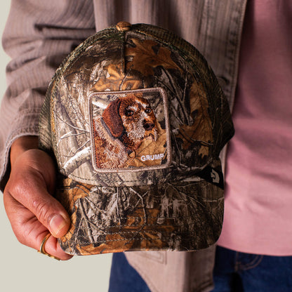 A person holds the Goorin Bros. Realtree Edge Grump Dog cap, showcasing a camo pattern, dog patch, and GRUMP embroidery on the front—perfect for channeling true grump energy.