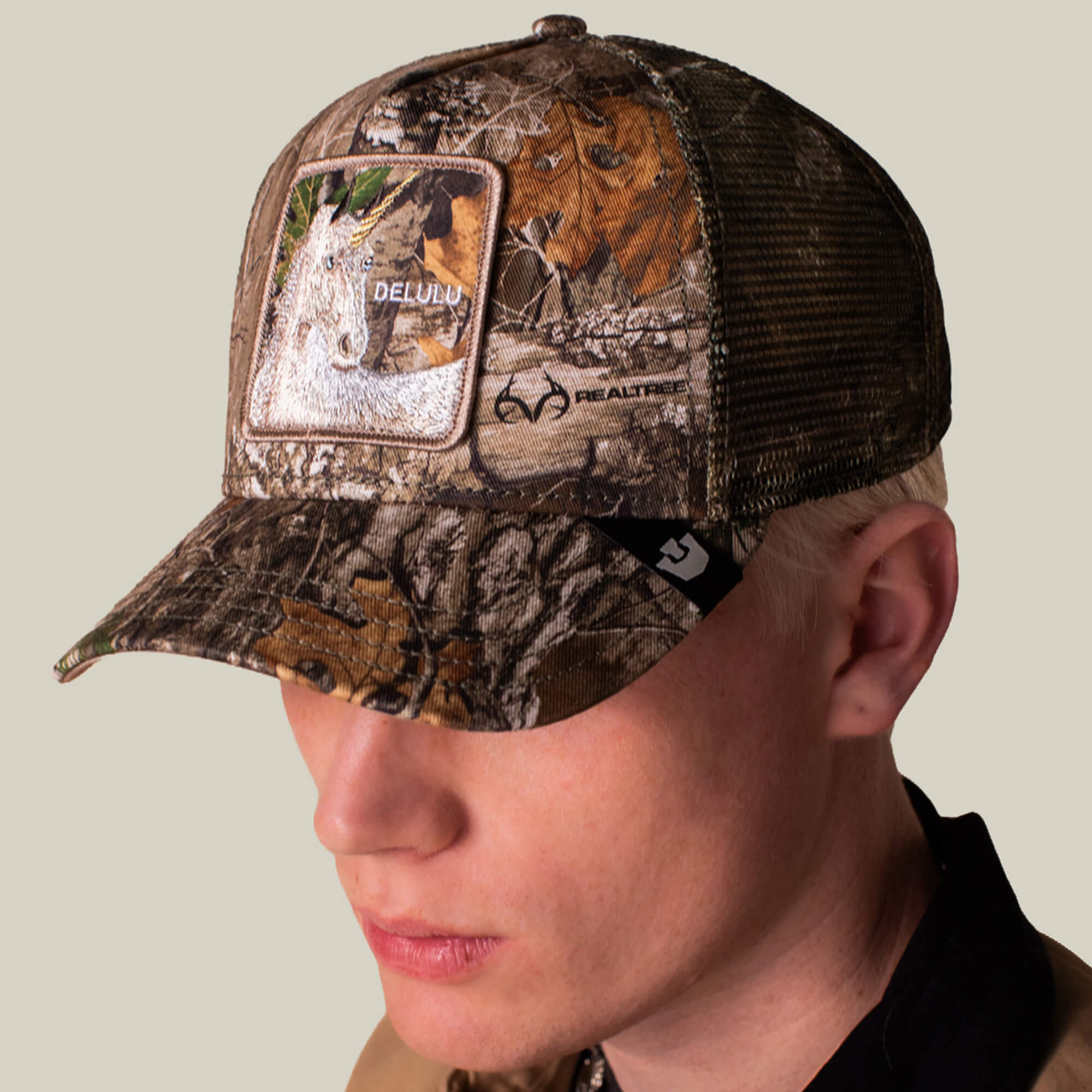 A person wears the Goorin Bros. Realtree Edge Delulu Unicorn camo trucker hat featuring a DELULU patch and Realtree logo, shown against a plain background.