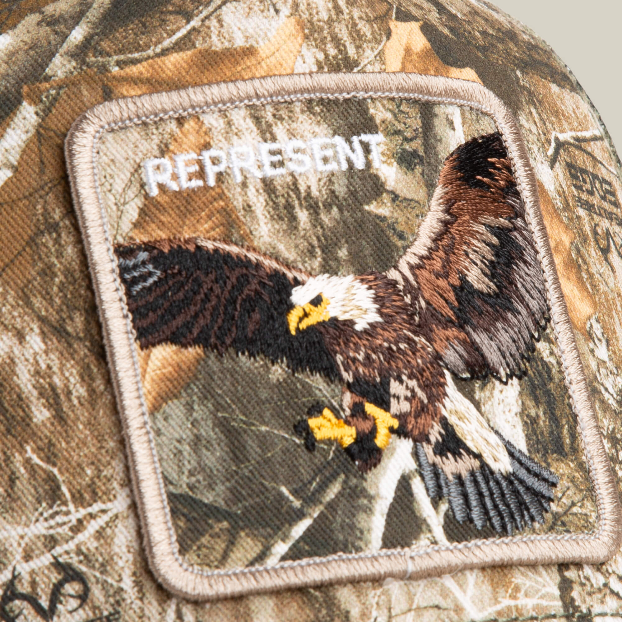 The Goorin Bros. Realtree Edge Represent Eagle patch showcases an embroidered bald eagle in flight with “REPRESENT” above, all set against a seamless Realtree camo colorway.