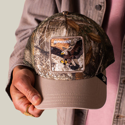 Someone in a beige jacket holds the Goorin Bros. Realtree Edge Represent Eagle trucker hat, featuring an embroidered eagle patch.