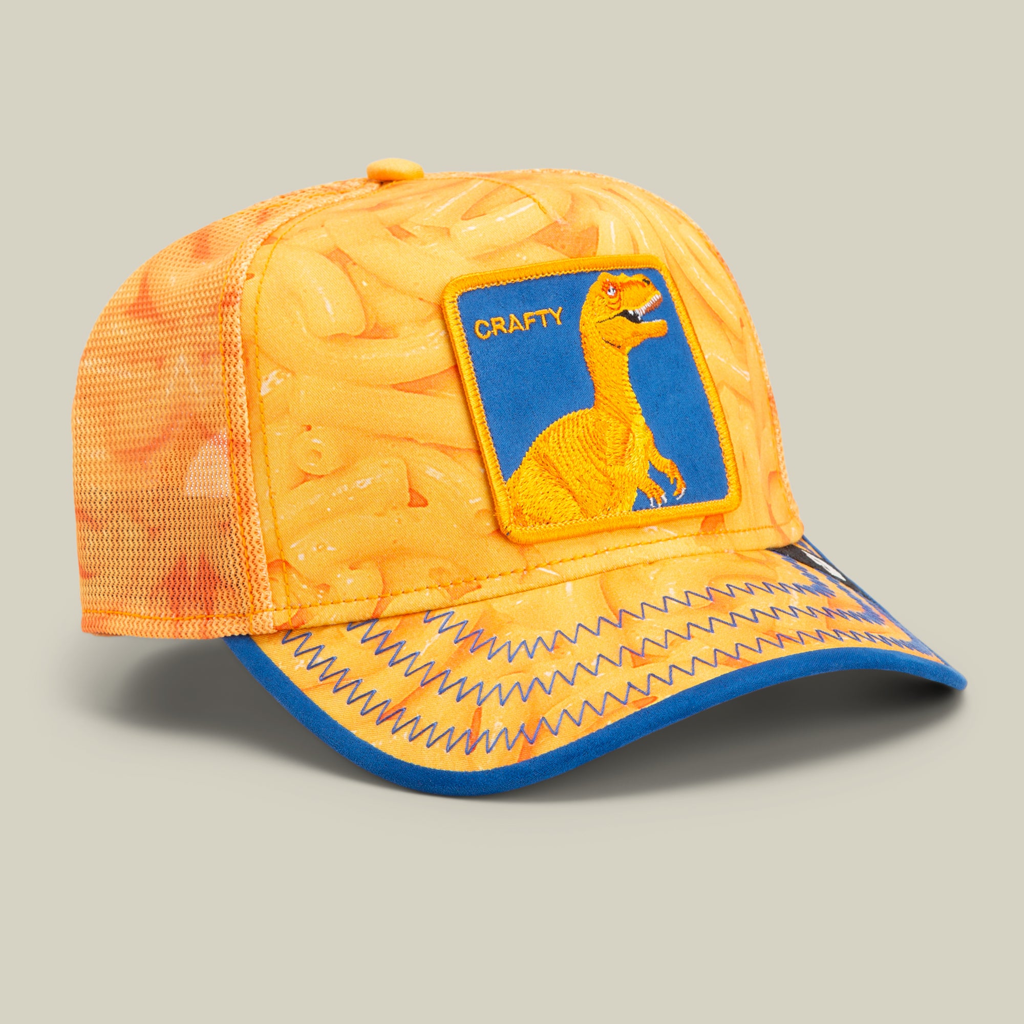 An orange mesh baseball cap with a macaroni pattern, blue zigzag stitching on the brim, and a patch featuring a yellow dinosaur labeled CRAFTY—perfect for fans of bold hat designs and unique trucker hats.