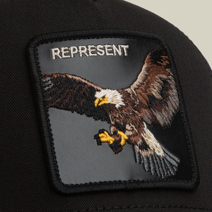 A close-up of the Represent Eagle Trucker by Goorin Bros., a black trucker hat with an embroidered eagle in flight and "REPRESENT" on the front patch.