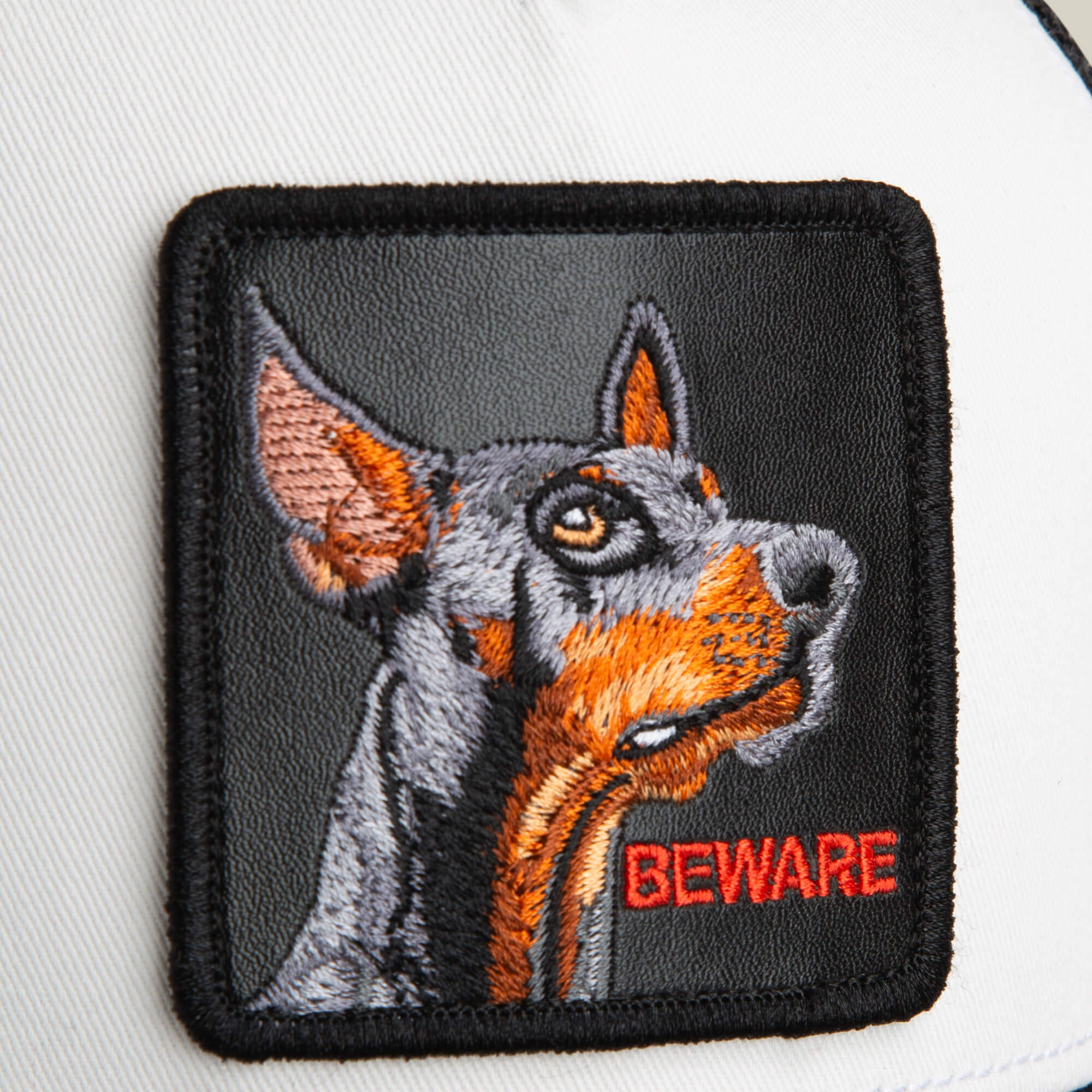 A close-up of The Beware Dog trucker hat by Goorin Bros., showing an embroidered Doberman patch with "BEWARE" in red—ideal for black or white trucker styles.