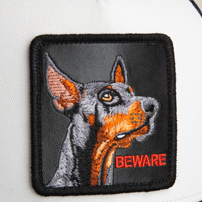 A close-up of The Beware Dog trucker hat by Goorin Bros., showing an embroidered Doberman patch with "BEWARE" in red—ideal for black or white trucker styles.