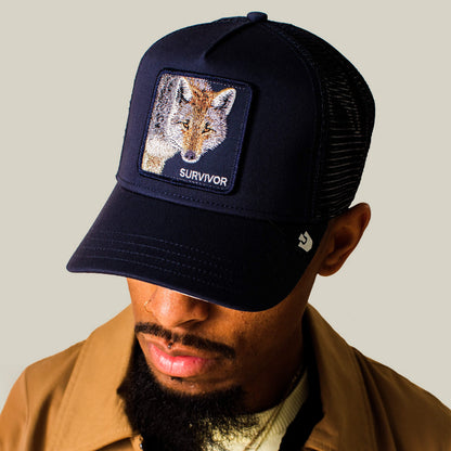 Man wearing the Goorin Bros. Mirage Survivor trucker hat featuring a fox patch labeled SURVIVOR, paired with a tan jacket and beige shirt, looking down.