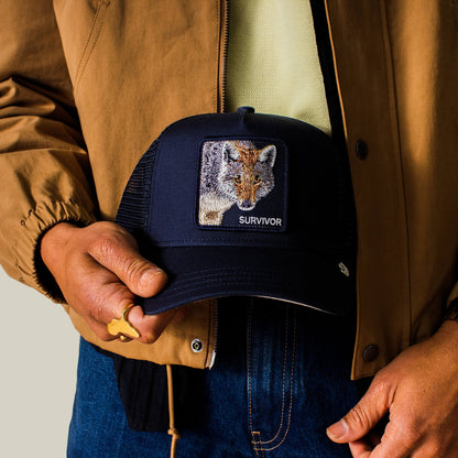 A person in a brown jacket holds a navy Goorin Bros. Mirage Survivor trucker hat featuring a fox patch labeled "SURVIVOR.
