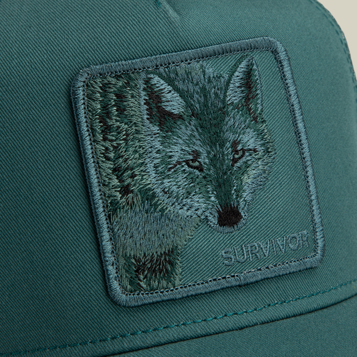 Dark teal Mirage Survivor trucker hat featuring a square patch embroidered with a wolf’s face.