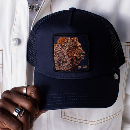 A person wearing a white jacket holds the Goorin Bros. Bear Trucker cap in navy, featuring a bear patch with the word "BEAR.