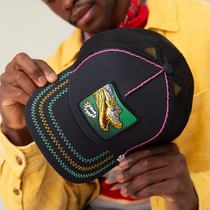 A person in a yellow jacket holds Goorin Bros.'s Crackle and Pop, Brother—a black mesh baseball cap with neon stitching and an embroidered gator patch—for a playful, bold accent.