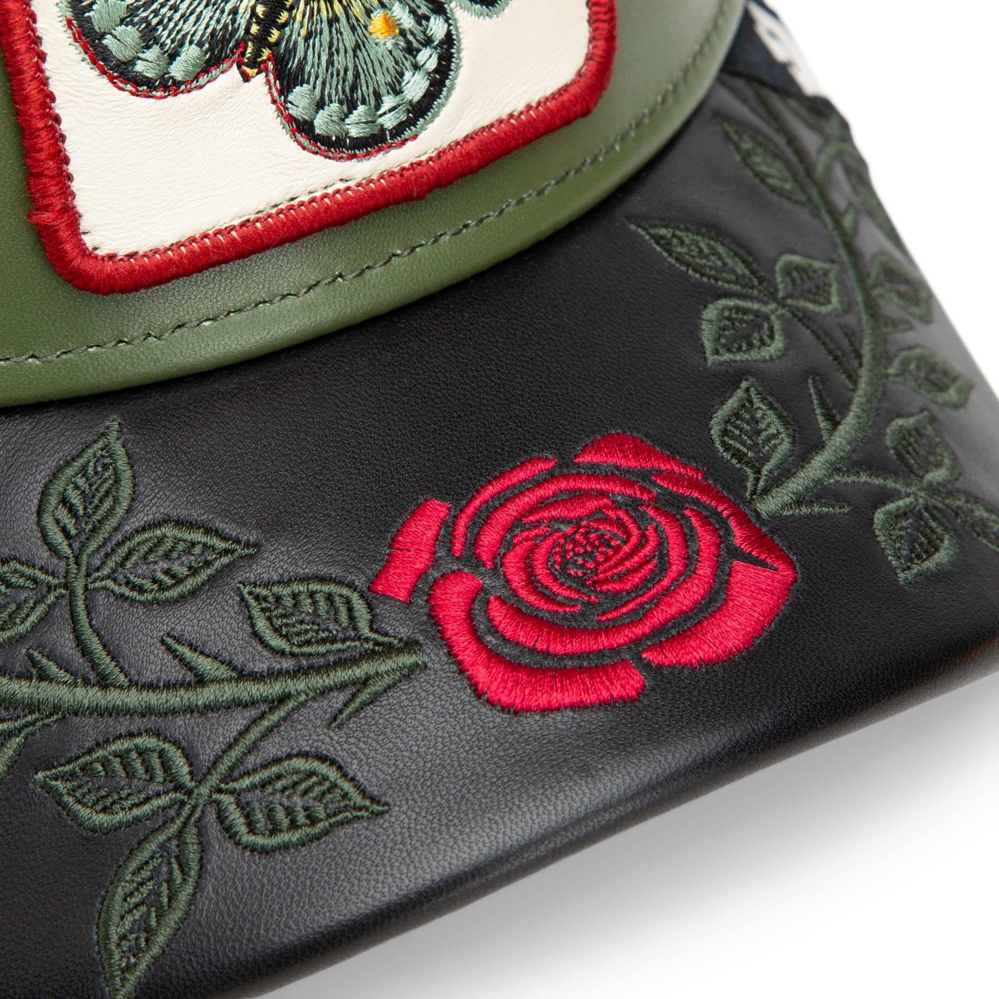A close-up of the Amor cap by Goorin Bros., in black and green, with embroidered green leaves, a red rose, and part of a butterfly patch—designs inspired by the elegance of AMOR jewelry.