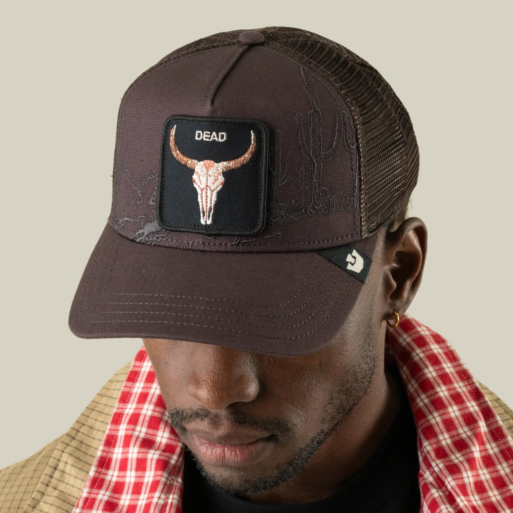 A man wearing the Goorin Bros. “Badlands Skull” dark mesh trucker hat featuring a patch with a bull skull and the word “DEAD,” paired with a checked red shirt under a tan jacket.
