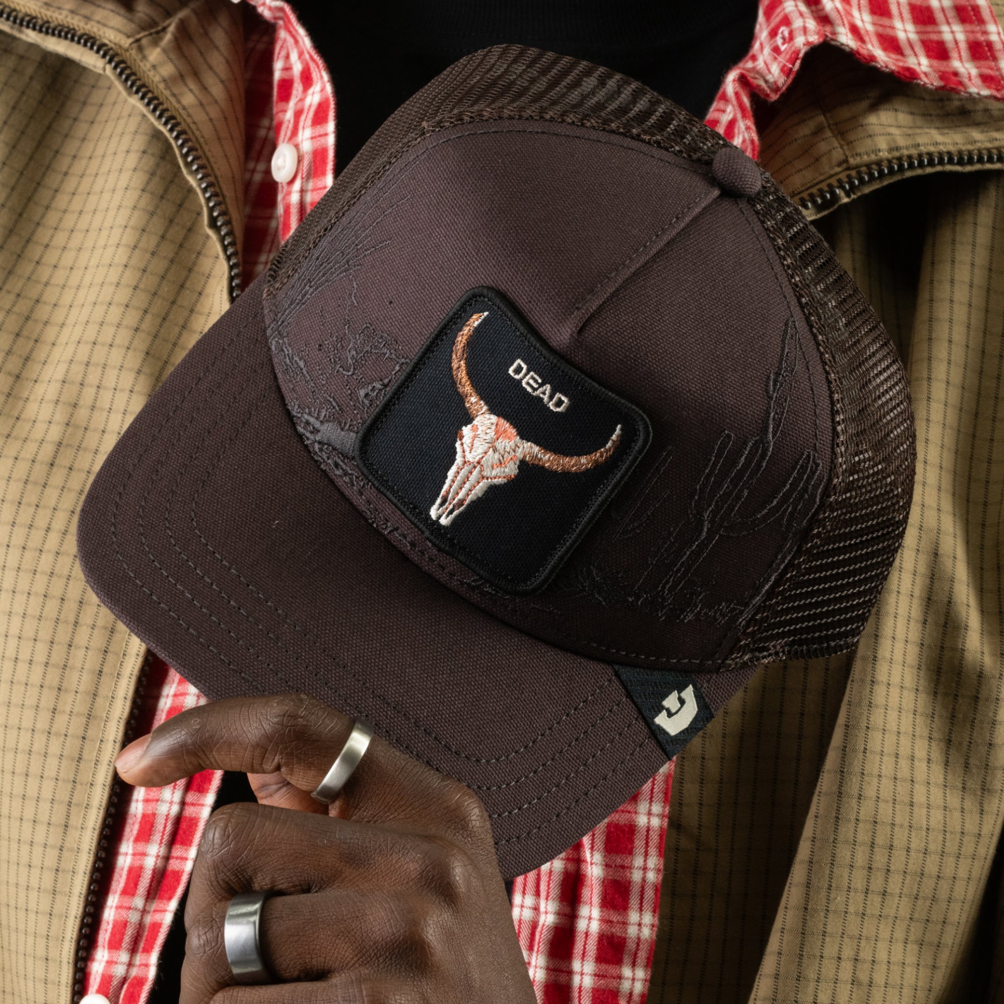A person holds the “Badlands Skull” trucker hat by Goorin Bros., a brown cap featuring a patch with a steer skull and the word DEAD. They are wearing a plaid shirt, tan jacket, and silver rings.