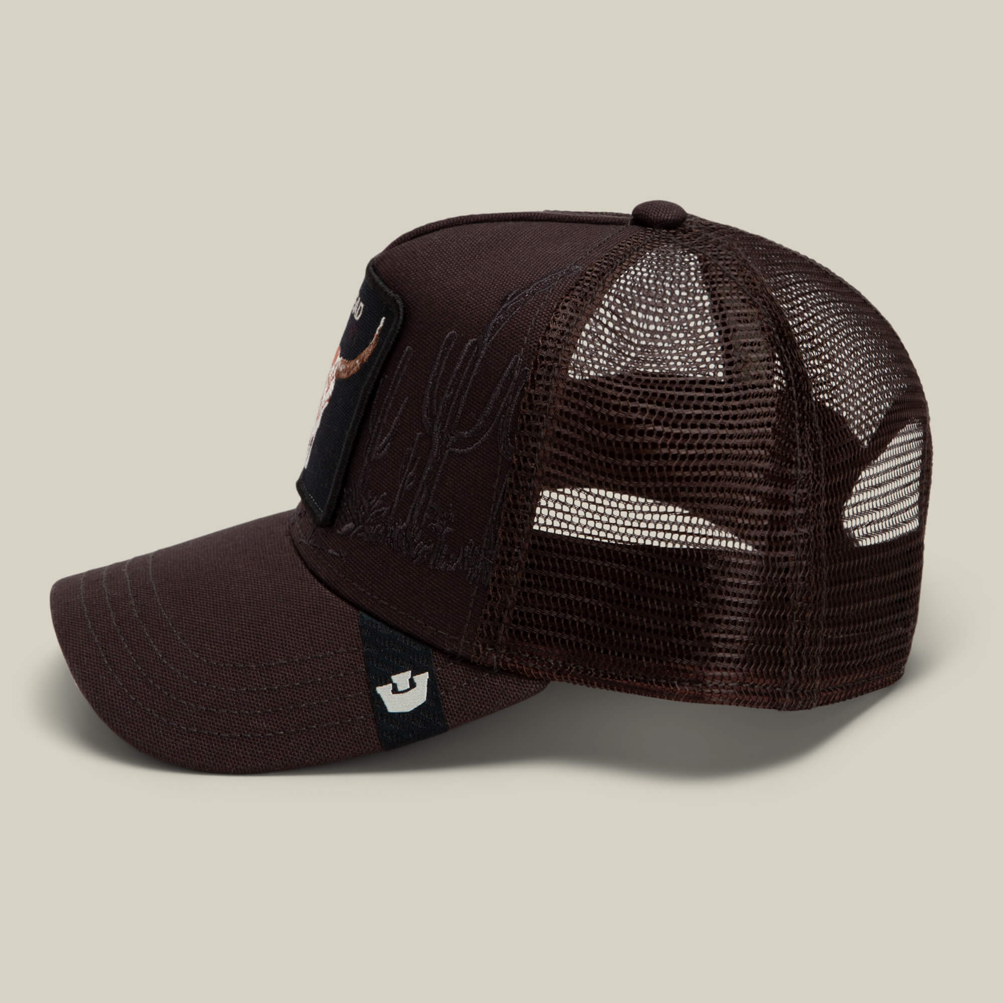The Goorin Bros. "Badlands Skull" brown mesh trucker hat features a curved bill, fabric front panel, and a patch on the front; shown from the side against a plain beige background.
