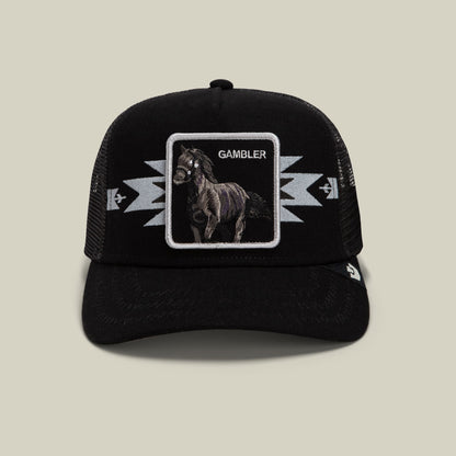 The Goorin Bros. Canyon Gambler black trucker hat features a mesh back, embroidered horse patch with "Gambler" text, and lightning bolt designs on the front panel, displayed against a neutral studio background.