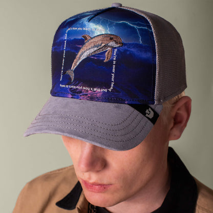 A person wearing the Goorin Bros. "Through the Storm - Dolphin" cap, which features a dolphin leaping from water amid lightning, with text curved around the dolphin.