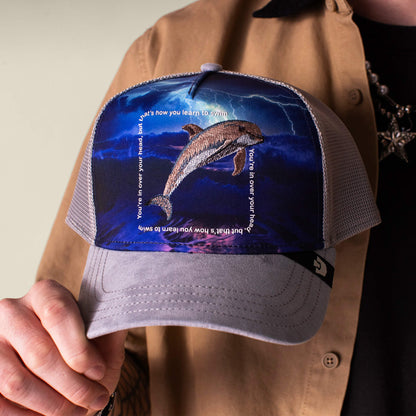 Someone holds the Goorin Bros. "Through the Storm - Dolphin" hat, featuring a gray dolphin graphic and the quote, "You're in over your head, that's how you learn to swim," printed around the image.