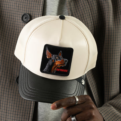 A person holds the Goorin Bros. Faux Leather Doberman trucker hat in beige and black, featuring a patch with a Doberman’s head and the word “BEWARE” in red letters.