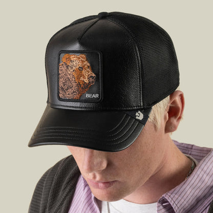 A person wearing the Goorin Bros. Faux Leather Bear trucker hat, featuring a black faux leather front with an embroidered grizzly patch, breathable mesh sides, a leather brim, and a small logo tag on the side, looking down.