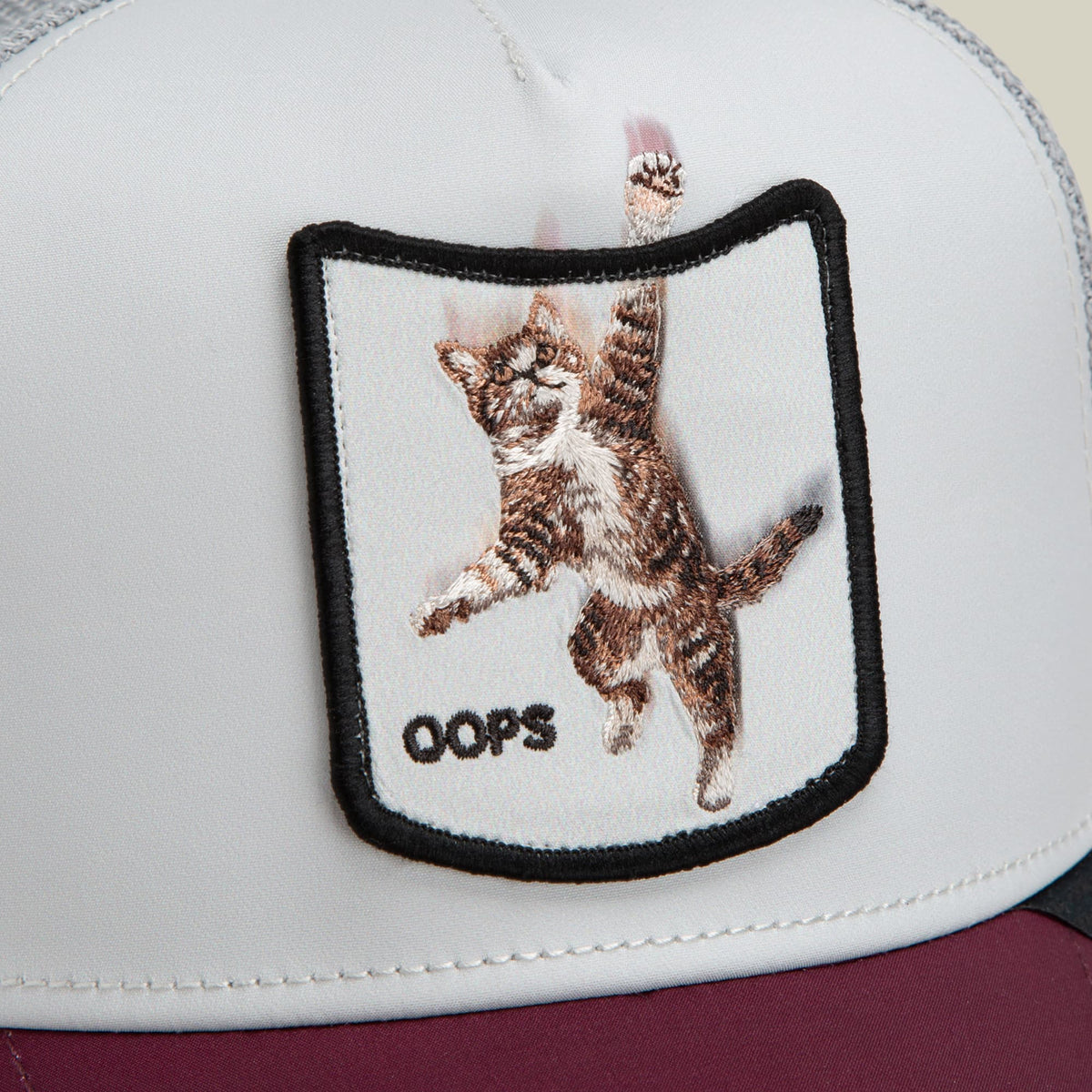 The Goorin Bros. Migration Oops trucker hat features a white front panel and maroon back with an embroidered patch of a brown tabby cat reaching upward and the word "Oops" below it, shown in a close-up product shot.