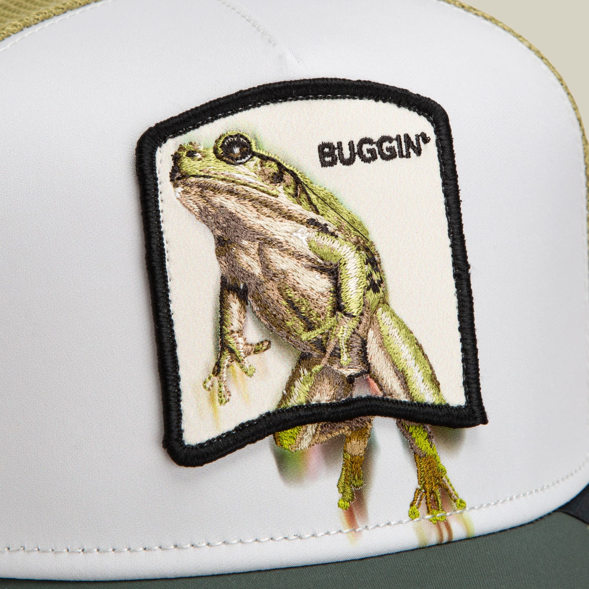 The Goorin Bros. Migration - Buggin' trucker hat features a white crown with black mesh back and a detailed embroidered green frog patch labeled "Buggin'" on the front.