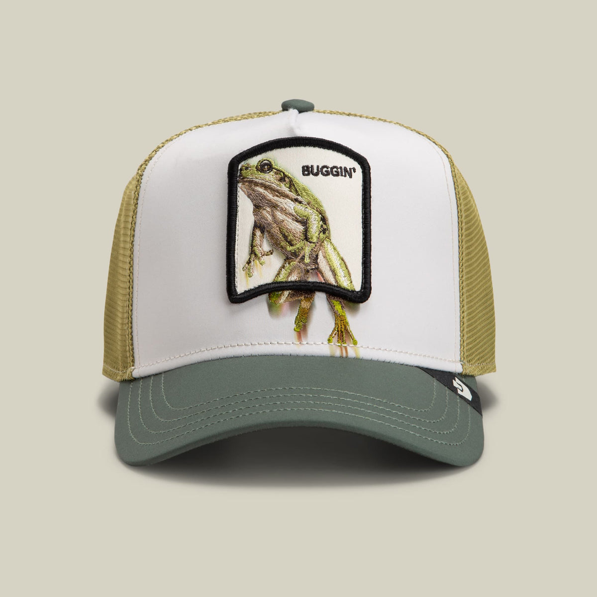 The Goorin Bros. Migration Buggin trucker hat features a white front panel with an embroidered frog patch labeled "Buggin'," mesh side panels in beige, and a sage green visor, displayed against a neutral background.