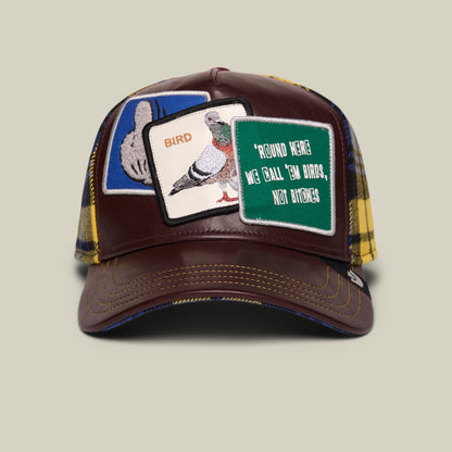 The Bird! by Goorin Bros. is a brown trucker hat featuring embroidered bird and text patches on the front, with yellow and navy plaid fabric on the mesh panels, displayed against a neutral background.