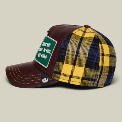 The Goorin Bros. Bird trucker hat features a brown leather front with yellow and navy plaid fabric back panel, a green patch with white text, and contrast stitching, displayed on a neutral background.