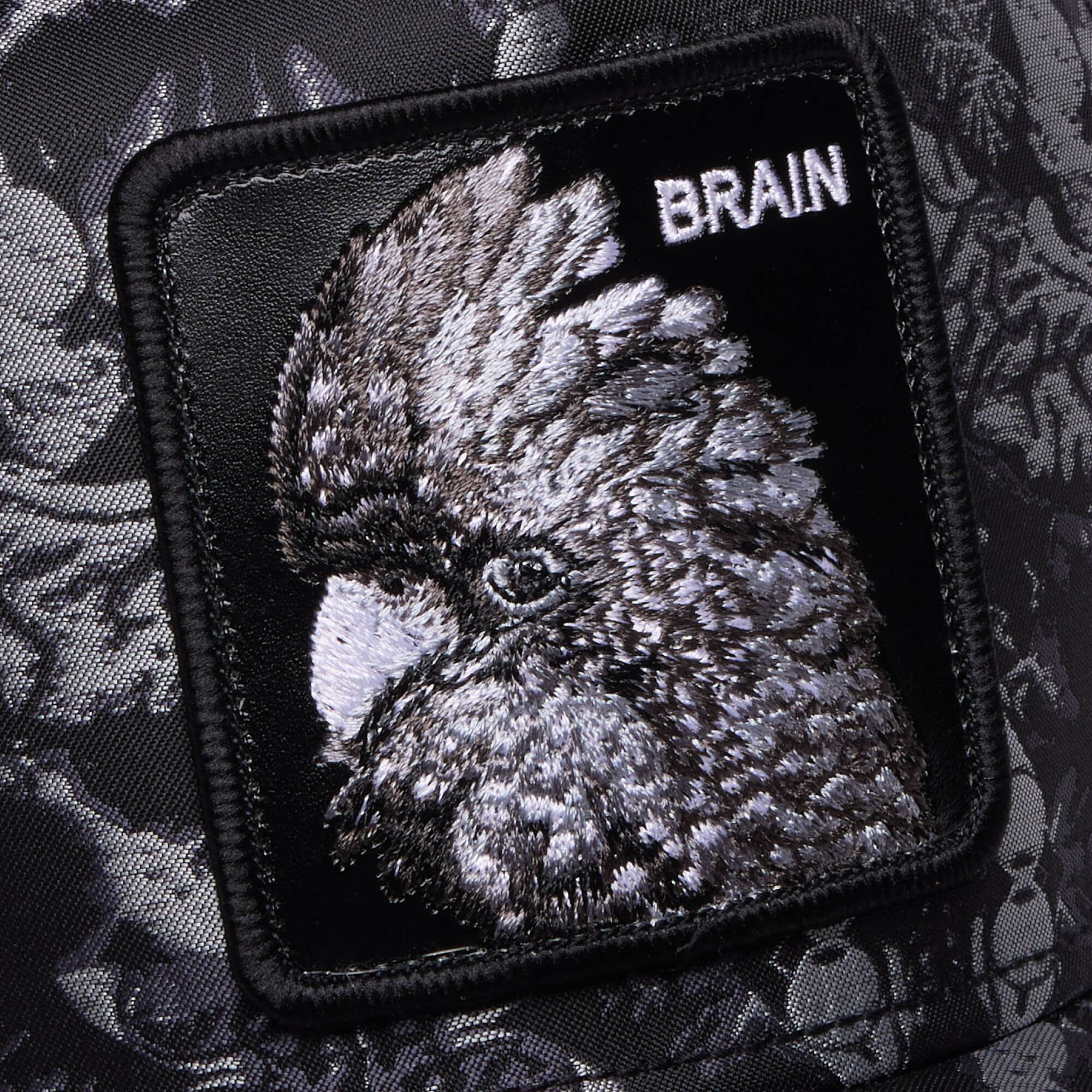 A close-up of the Goorin Bros. "Gray Matter" patch in black and grey, embroidered with a bird and the word BRAIN, set against patterned fabric—a stylish nod to intellect.