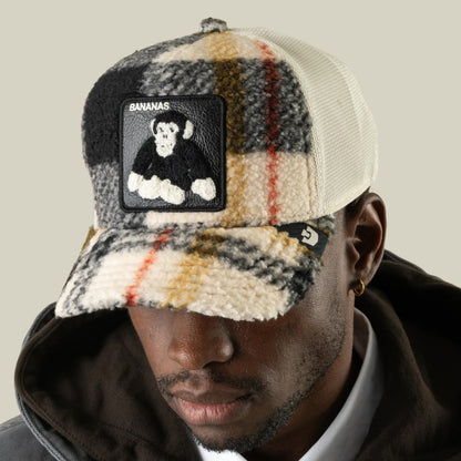A man wearing the Goorin Bros. Double Stuffed - Bananas plaid fleece snapback hat with a monkey patch, paired with a black hoodie and gold hoop earring, posed against a neutral background.